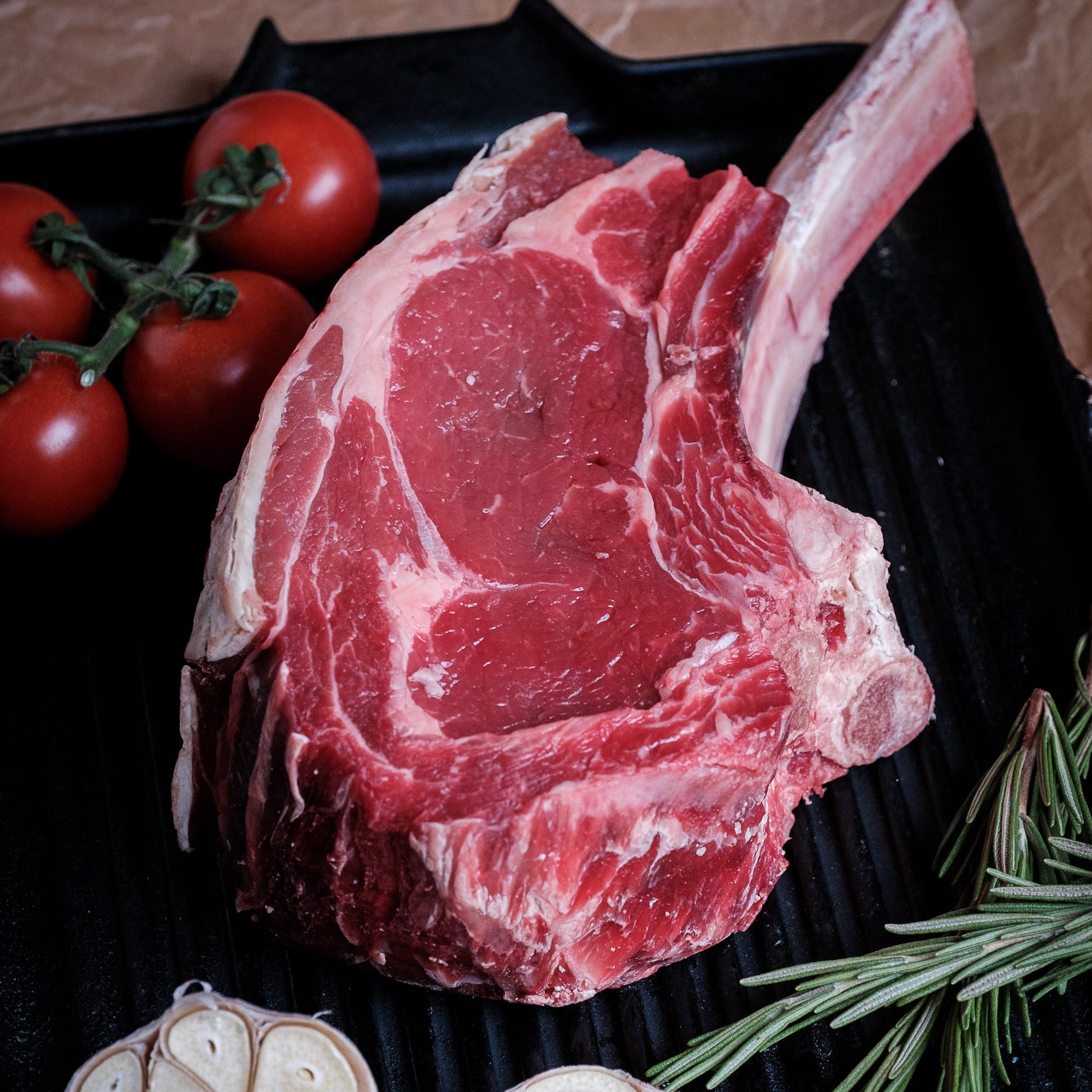 A raw tomahawk steak with a bone, displayed on a black grill plate with halved garlic bulbs, fresh rosemary sprigs, and red cherry tomatoes on a brown textured background.