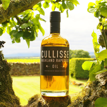 A bottle of Cullisse Highland Rapeseed Oil stands on a mossy tree branch outdoors, framed by green leaves, with a blurred garden and stone wall in the background.