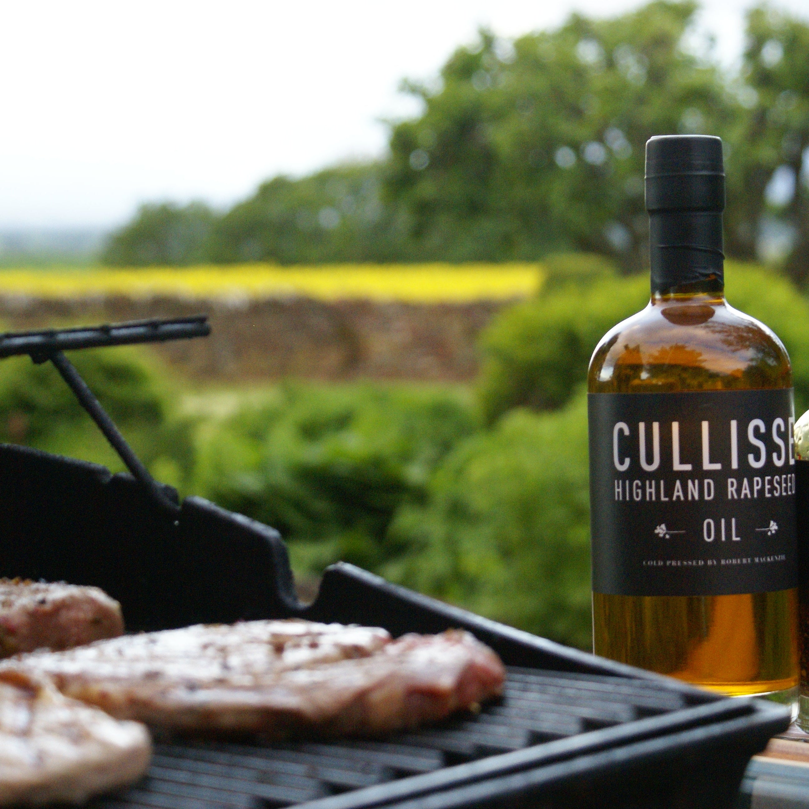 Bottle of Cullisse Highland Rasper Oil and Marinate seasoning oil displayed next to a grill with steaks, set against a blurred outdoor Scottish landscape.