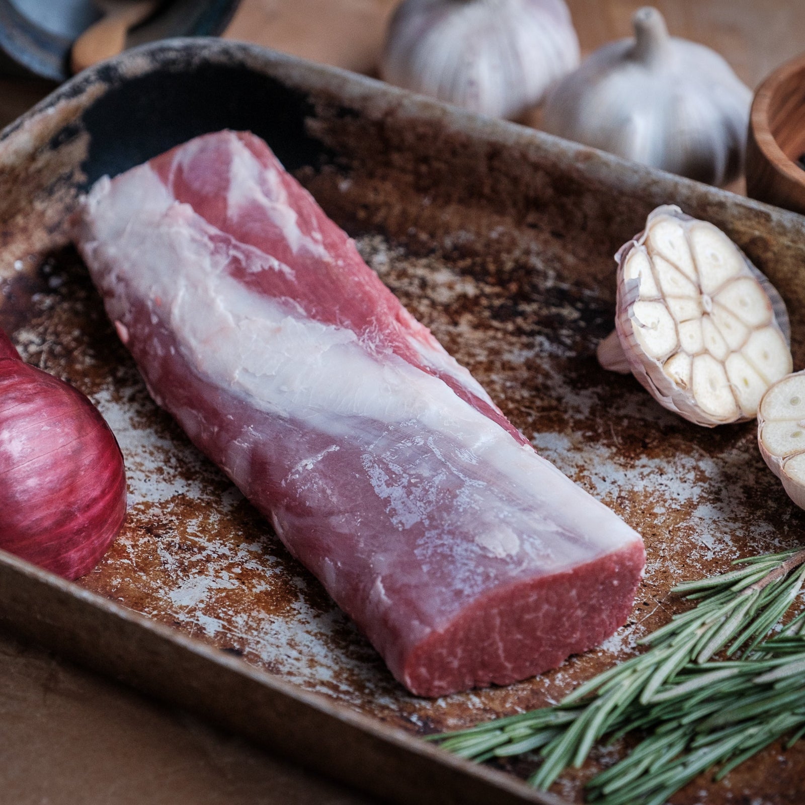 Raw canon of lamb on a baking tray with garlic, rosemary, and red onions.
