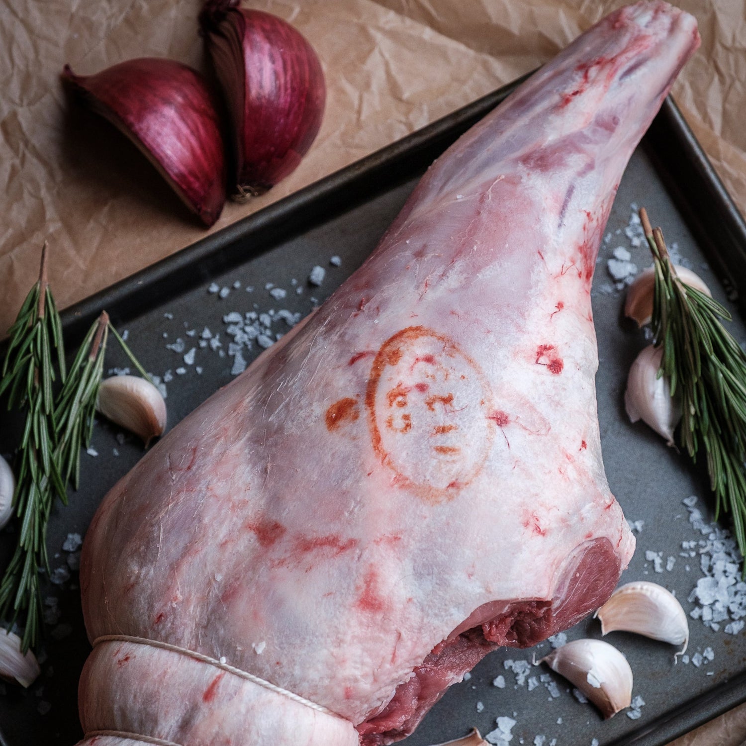 Raw carvery leg of lamb on a baking tray with garlic, rosemary, and a bowl of peppercorns.