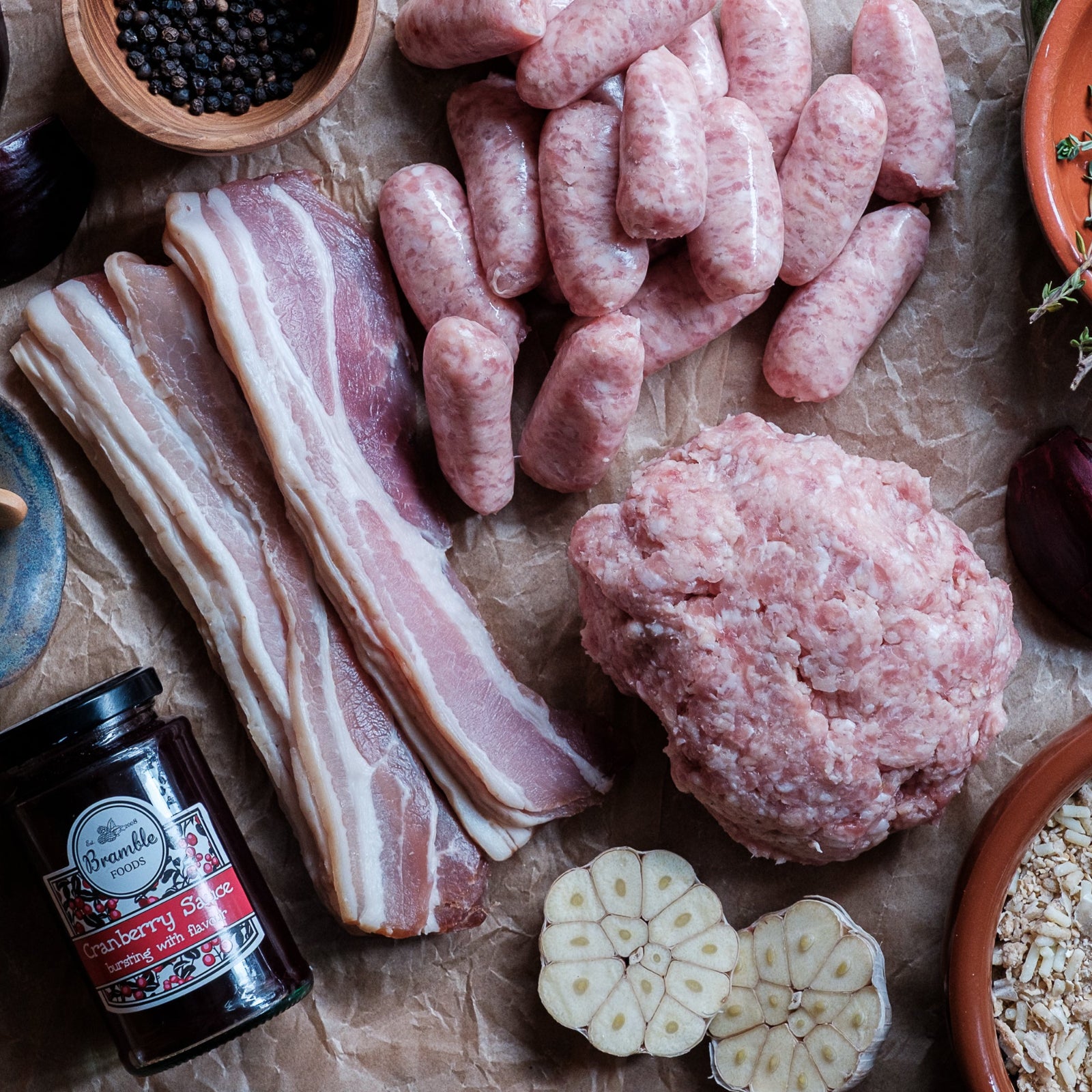 Raw meat products including sausages, bacon, and ground meat on a wooden surface with herbs and spices.