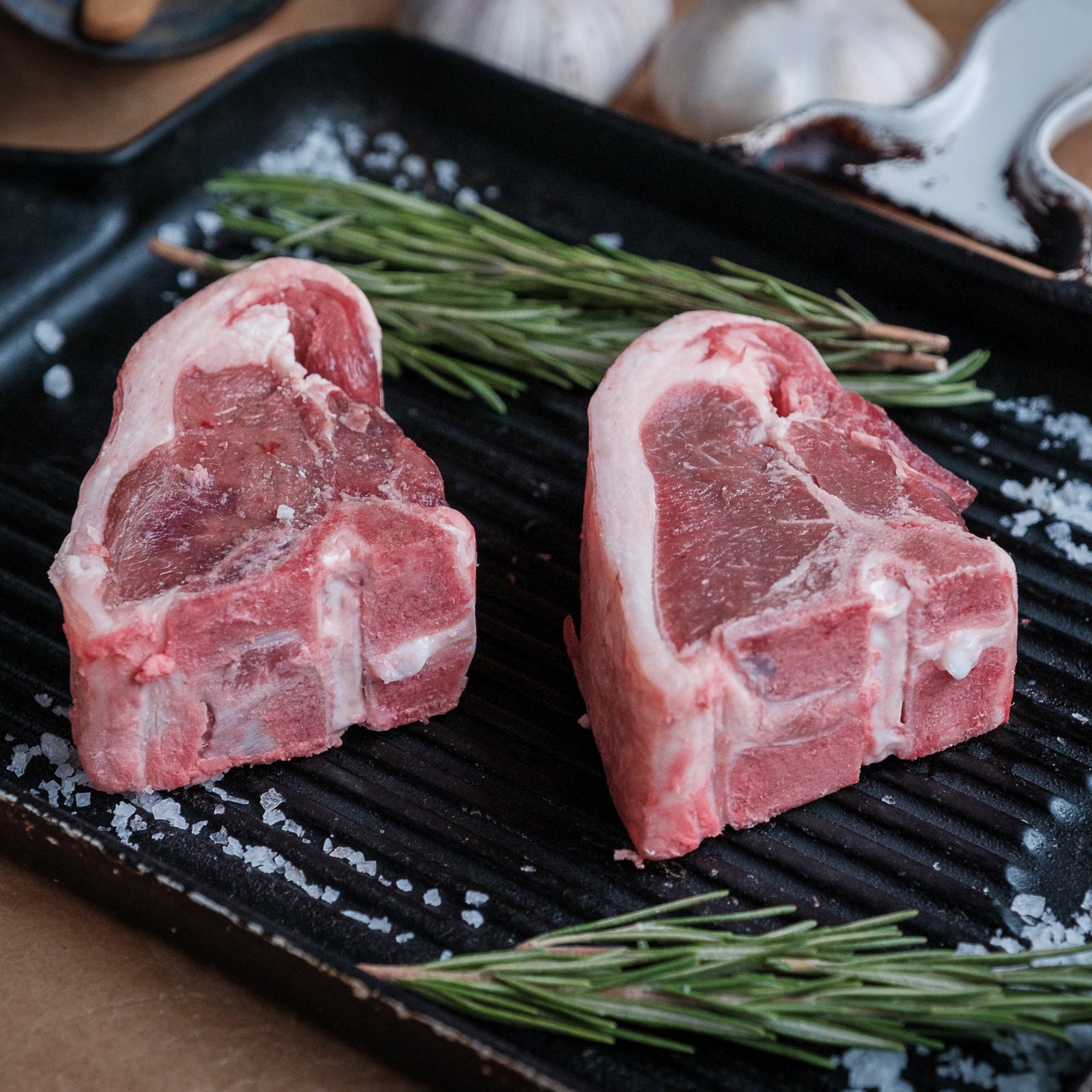Two raw lamb chops on a grill pan with rosemary and salt.
