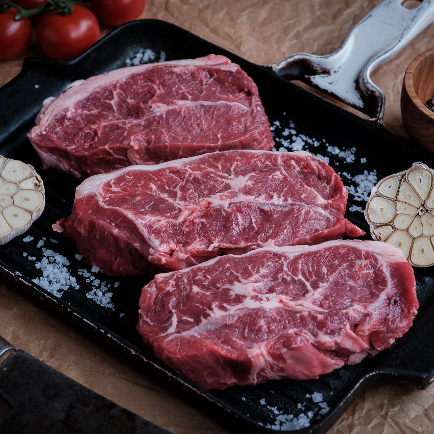 Three raw feather steaks on a black cast iron skillet with garlic, tomatoes, and a wooden bowl of peppercorns on a rustic background.