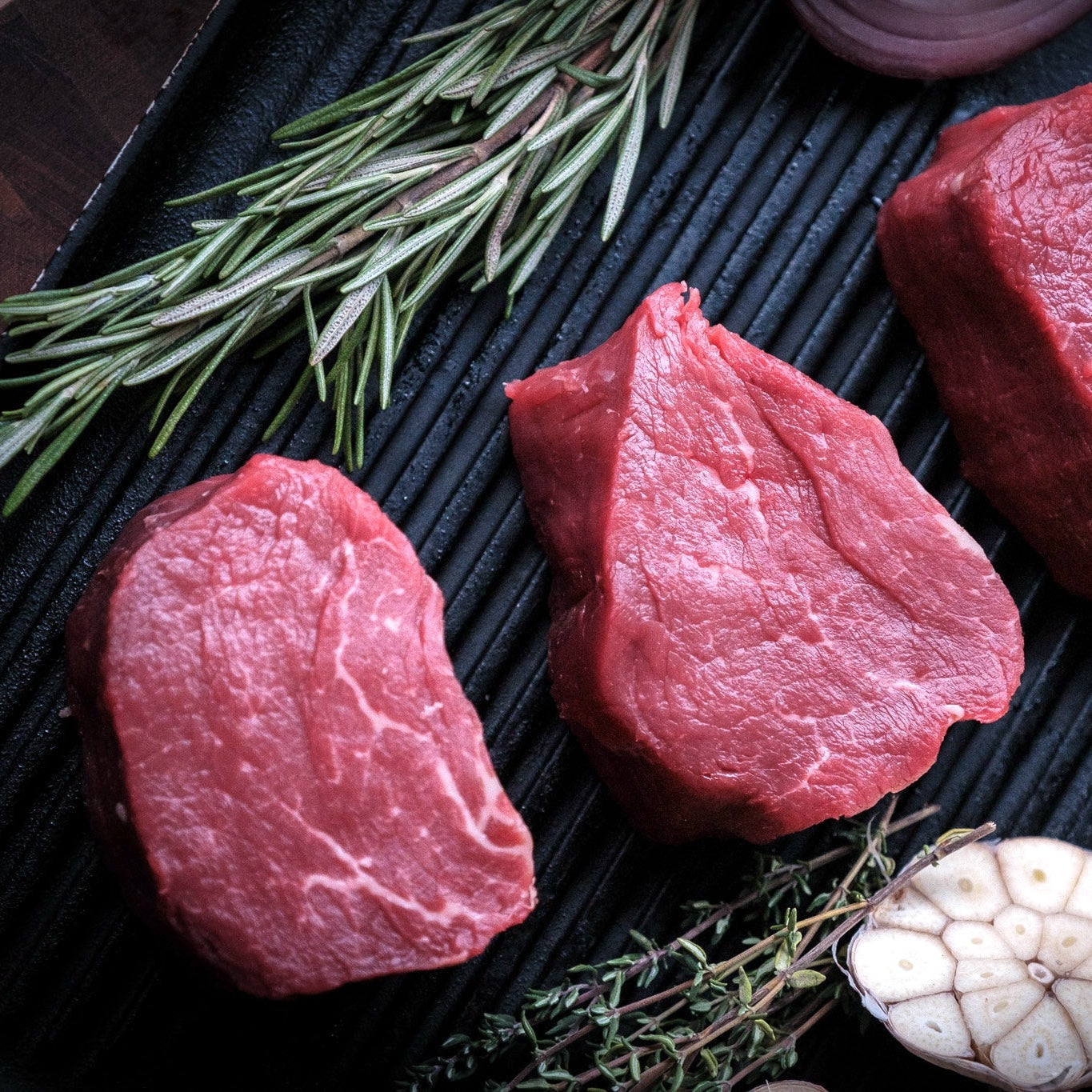 Three raw 50 days aged beef fillet steaks on a black grill pan with rosemary, garlic, and oil, photographed from above.