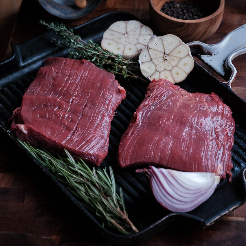 Two raw flank steaks on a grill pan with herbs and vegetables on a wooden surface