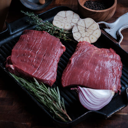 Two raw flank steaks on a grill pan with herbs and vegetables on a wooden surface