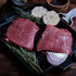 Two raw flank steaks on a grill pan with herbs and vegetables on a wooden surface