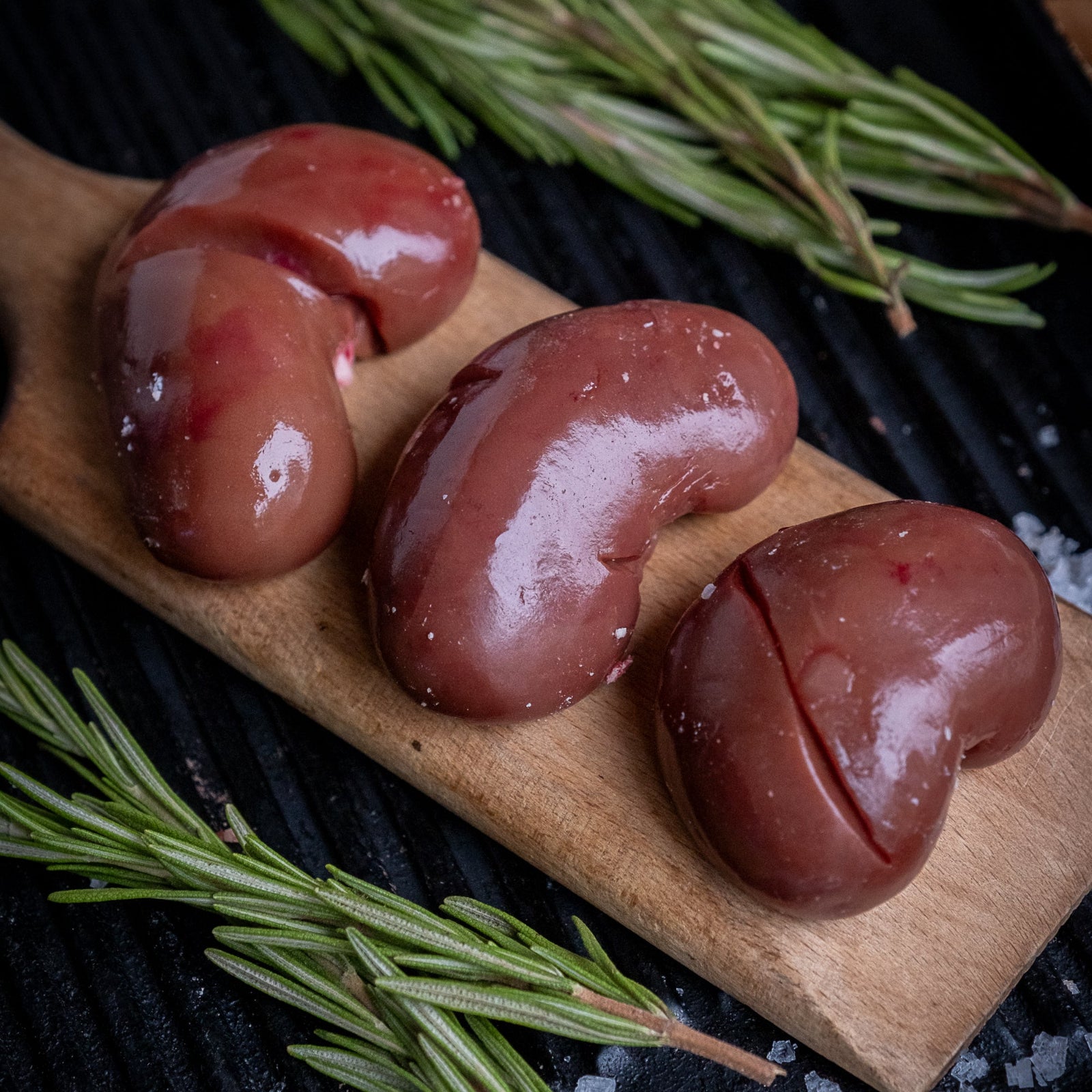 Three raw lamb kidneys on a wooden board with rosemary and salt crystals.