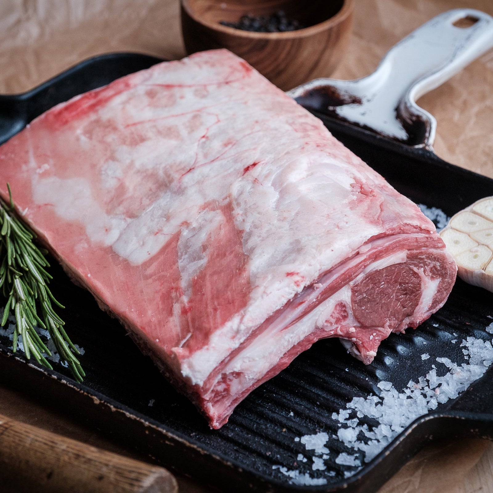 Raw lamb loin on a black grill pan with herbs and garlic on a wooden surface
