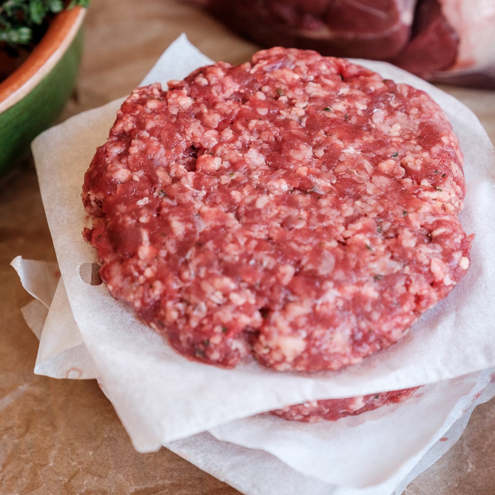 Raw burger patty on a piece of paper with fresh herbs and a tomato in the background.