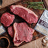 Raw steaks on a wooden cutting board with Potts seasoning packets and herbs.