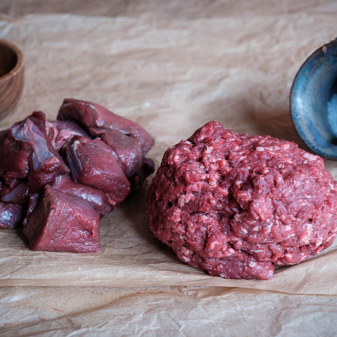 Raw diced and minced venison on brown paper with a wooden bowl and blue container.