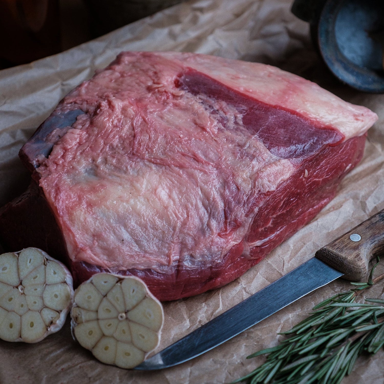 Raw plate of beef on brown paper with garlic, rosemary, and a knife.