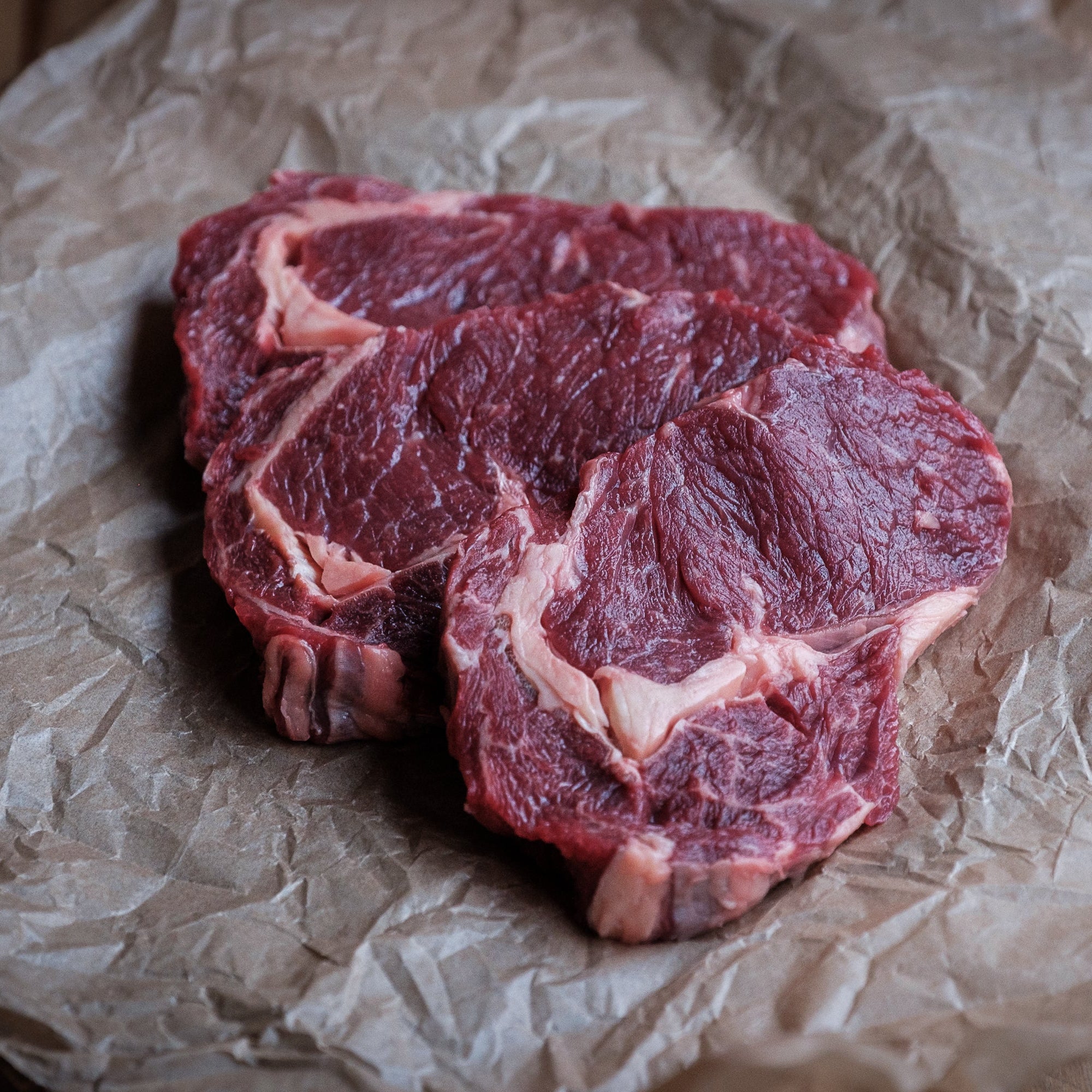 Three raw 50 day aged rib eye steaks with visible marbling, resting on crumpled brown butcher paper over a dark wooden surface.
