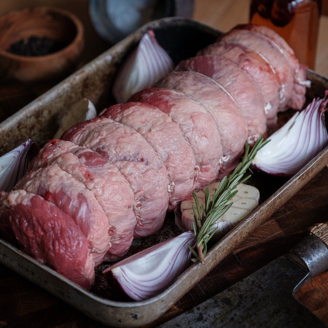 Raw rolled beef brisket with onions and rosemary in a roasting pan on a wooden surface