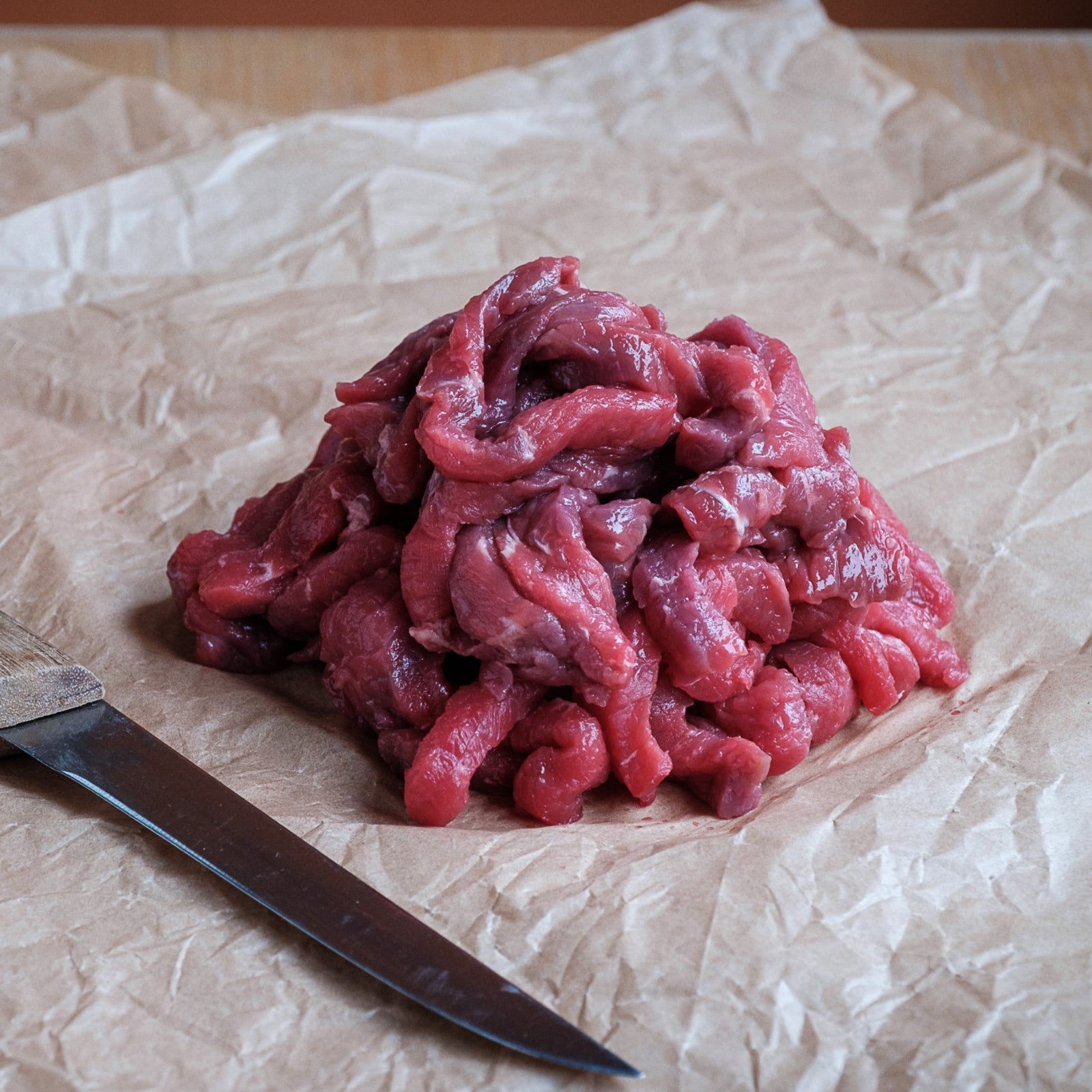 Raw beef stir fry strips and a knife on crumpled paper with a wooden surface.
