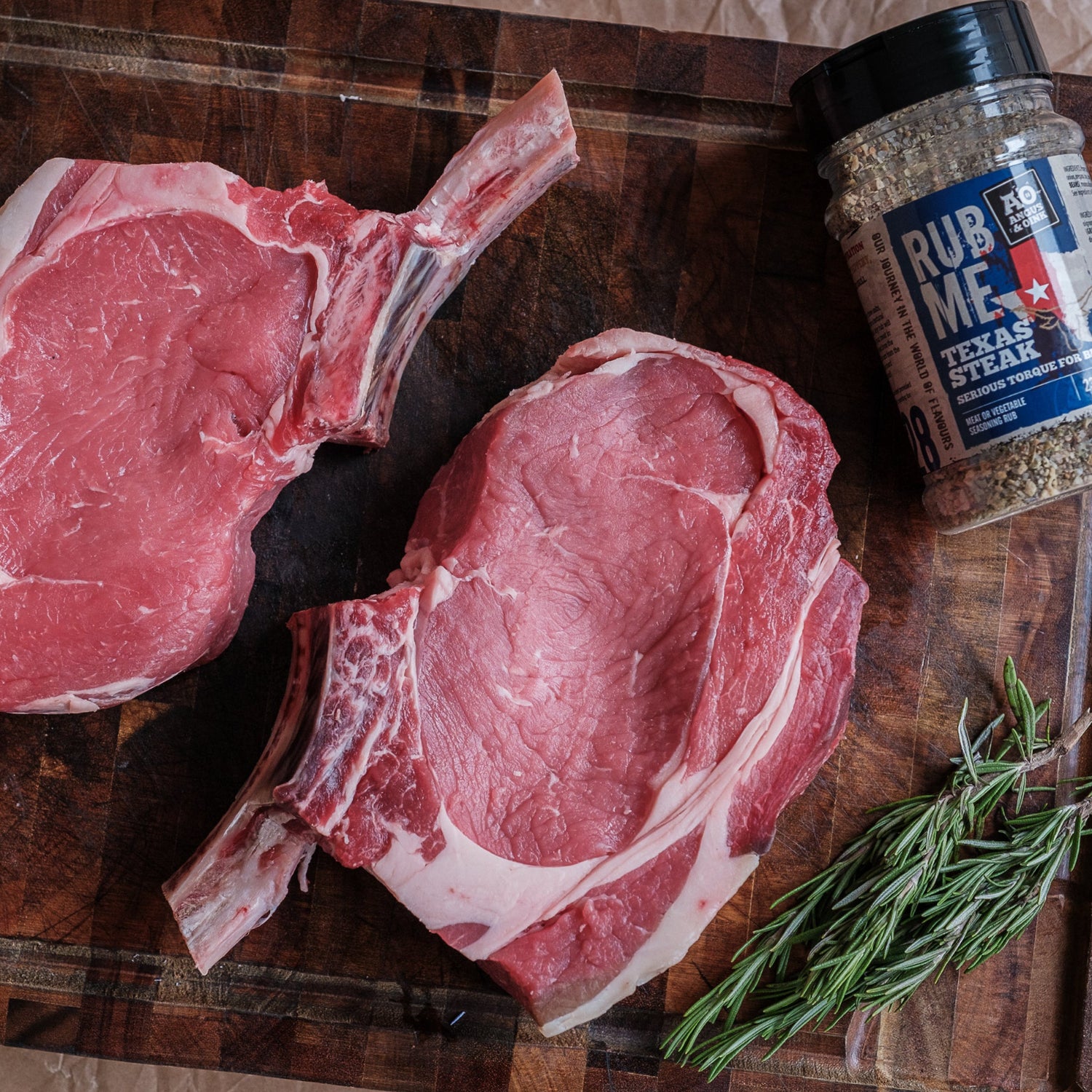 Two raw tomahawk steaks on a wooden cutting board with a jar of seasoning and rosemary.