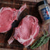 Two raw tomahawk steaks on a wooden cutting board with a jar of seasoning and rosemary.