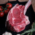 A raw tomahawk steak with a bone, displayed on a black grill plate with halved garlic bulbs, fresh rosemary sprigs, and red cherry tomatoes on a brown textured background.